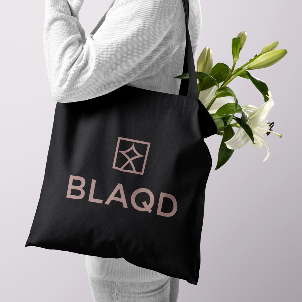 The BLAQD stacked lockup logo in rose on a midnight tote bag filled with flowers and being carried over someones shoulder